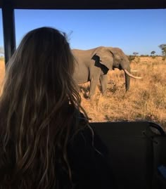 an elephant walking across a dry grass covered field next to a woman in a black jacket