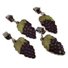 three grapes are hanging from the side of a pair of silver earrings on a white background