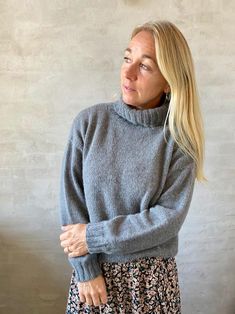 a woman wearing a gray sweater and floral print skirt standing in front of a white wall