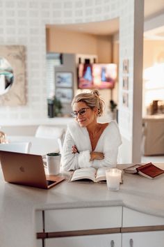 a woman sitting at a table with a laptop and coffee in front of her, reading a book