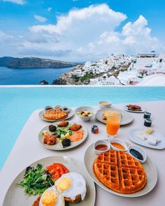 a table with plates of food on it next to a pool and the ocean in the background