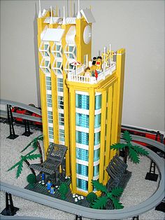 a lego model of a yellow building with palm trees on the ground and cars in front