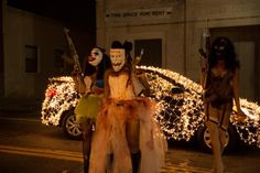 three women dressed in costumes standing next to a car with christmas lights all around them