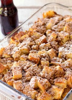 a casserole dish with bread and cinnamon toppings in a glass baking dish next to a bottle of syrup