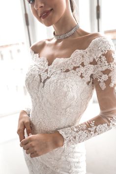 a woman wearing a white wedding dress with long sleeves and lace on the shoulders, holding her hand in her pocket