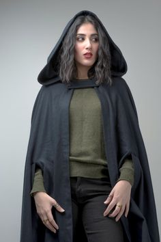 a woman wearing a black cloak and green shirt with her hands on her hips, standing in front of a gray background