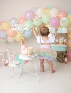 a baby girl standing in front of a cake with balloons behind her and a birthday sign above it