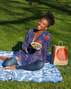 a woman sitting on the grass with a bowl of food in her hand and shopping bag