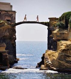 two people are walking on a bridge over the water near some rocks and cliffs that look like they have been built into the ocean