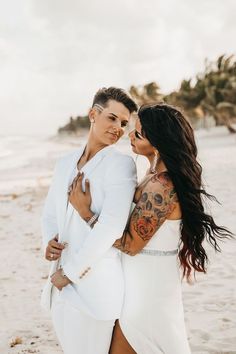 two women in white dresses standing on the beach with their arms around each other and looking into each others eyes