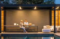 a man and woman sitting on a couch next to a swimming pool in front of a house