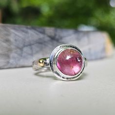 This tourmaline cabochon has a wonderful pink coloring with lots of interest and layers of depth. I set it in a bezel on a domed band with a high karat gold accent.  This smooth domed tourmaline is set in a fine silver bezel on a sterling silver backplate. The band is a half round domed shape that has been hammered to give it texture. A luxurious ball of solid 18K gold have been soldered to the band as an accent. The ring is marked with my makers mark, 925 and 18K. It is lightly oxidized to highlight the texture and the gold accents. It is a size 7 1/4. I was happy with how this ring evolved. It is a stunning ring with lots of panache! I hope you like it! Pink Round Tourmaline Jewelry, Pink Round Cabochon Ruby Ring, Pink Cabochon Ruby Ring, Pink Tourmaline Sapphire Ring, Pink Cabochon Ruby Ring Gift, Tourmaline Bezel Set Ring In Oval Cabochon, Tourmaline Ring With Bezel Setting In Oval Cabochon, Tourmaline Bezel-set Oval Cabochon Ring, Unique Round Cabochon Ruby Ring