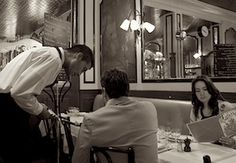 people sitting at tables in a restaurant with menus on the table and two waiters standing over them