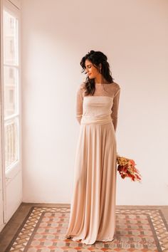 a woman in a long dress is standing by a window and holding a flower bouquet