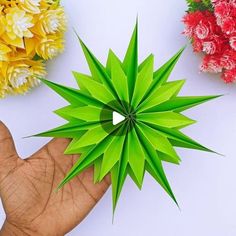 a hand holding a green paper flower in front of three different colored flowers on a white surface