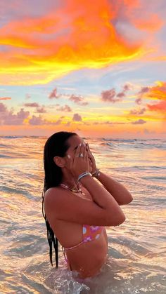 a woman standing in the ocean while talking on her cell phone with an orange and pink sunset behind her