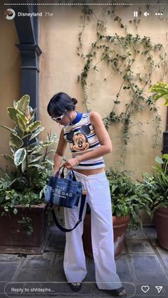 a woman holding a mickey mouse bag in front of some potted plants and greenery