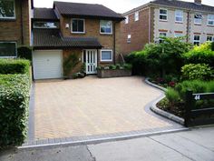 a brick driveway in front of two houses with bushes and shrubs on either side of the driveway