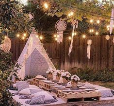 an outdoor dining area with lights strung over the table and pillows on the ground, in front of a wooden fence