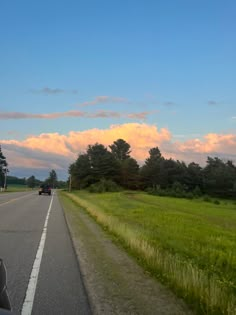 a car is driving down the road in front of some trees and grass at sunset