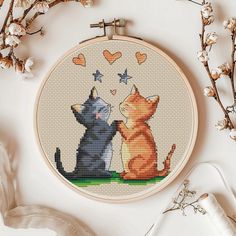 two cats sitting next to each other in front of a cross - stitch hoop with hearts on it