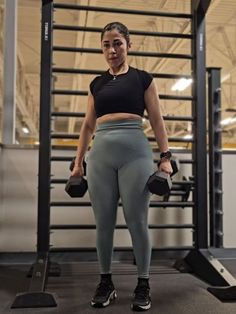 a woman standing in front of a gym machine holding two kettles and posing for the camera