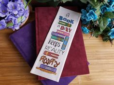 a cross - stitch bookmark on top of some purple and red cloths next to blue flowers