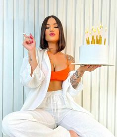 a woman holding a cake with candles on it in front of her face and wearing white pants