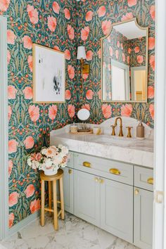 a bathroom with floral wallpaper and marble counter tops, gold faucet, vanity mirror, and stool