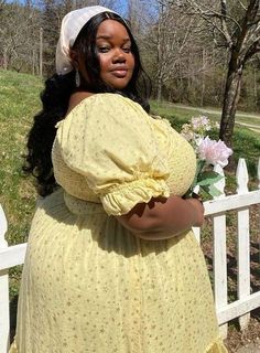 a woman in a yellow dress is standing by a white fence and holding a flower