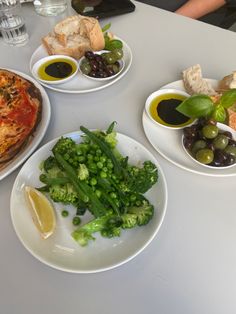 three plates of food on a table with olives, broccoli and bread