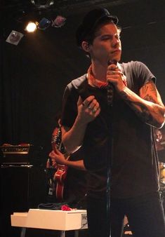 a man standing in front of a microphone on stage with other people behind him and one person holding a guitar