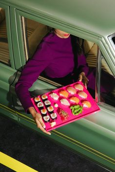 a woman holding a pink tray with sushi on it in front of a green car