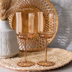 two wine glasses sitting on top of a woven place mat next to a white vase