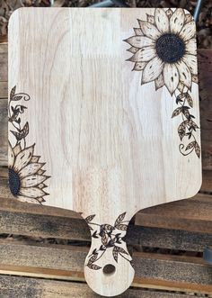 a wooden cutting board with sunflowers and leaves painted on the side, sitting on top of wood planks