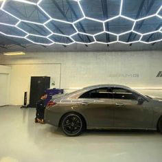 a man standing next to a silver car in a garage with lights on the ceiling