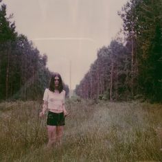 a woman standing in the middle of a field with trees behind her and grass all around her