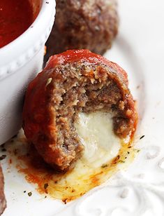 some meatballs and sauce on a white plate with a cup of tomato soup in the background