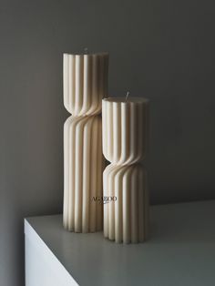 two tall white candles sitting on top of a table next to each other in front of a gray wall