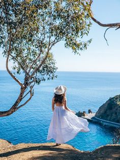 a woman in a white dress and hat looking out at the ocean from a cliff