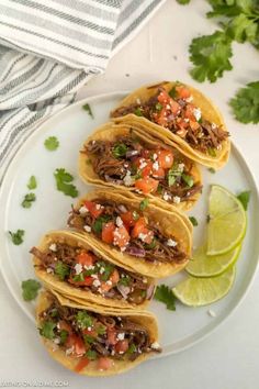 three shredded beef tacos on a white plate with limes and cilantro