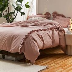 a bed with pink comforter and pillows on it next to a plant in a vase