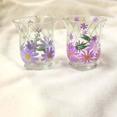 two glass vases sitting on top of a white cloth covered tablecloth with purple and yellow flowers painted on them