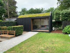 a small black building with a green roof and wooden benches in the front yard, surrounded by greenery