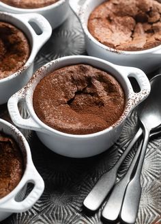 four white dishes filled with chocolate pudding on top of a silver tray next to spoons