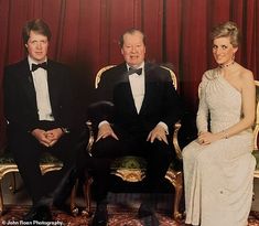 two men and a woman in formal wear sitting on gold chairs with red curtains behind them