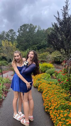 two young women are posing for the camera in front of colorful flowers and trees on a cloudy day