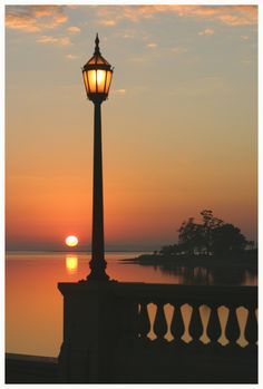 the sun is setting behind a lamp post in front of a body of water with trees