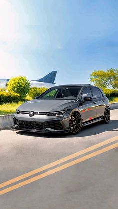 a gray car driving down the road next to an airplane on a runway with grass and trees in the background