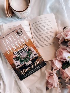 an open book sitting on top of a bed next to pink flowers and a cup